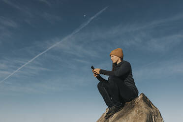 Frau mit Smartphone sitzt auf einem Felsen vor blauem Himmel - DMGF00932