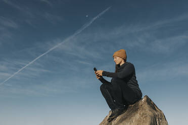 Woman using smart phone sitting on rock in front of blue sky - DMGF00932