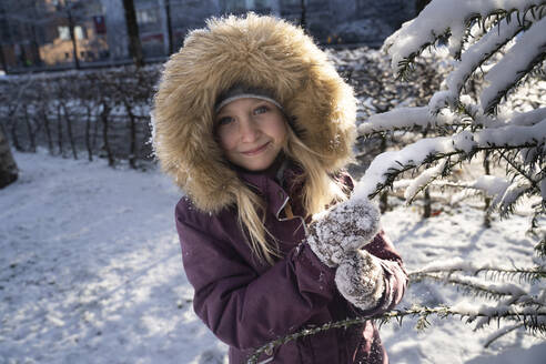 Lächelndes Mädchen mit Pelzmütze an einem Tannenbaum - NJAF00164