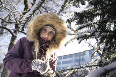 Lächelndes Mädchen mit Pelzmütze unter einem Tannenbaum stehend - NJAF00165