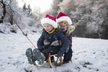 Smiling brother and sister sitting on sled in snow - NJAF00167