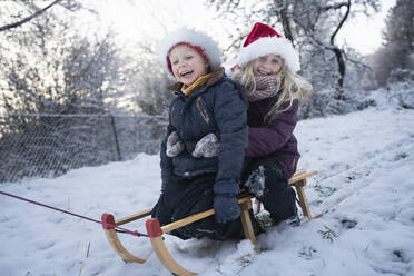 Glückliche Geschwister auf einem Schlitten im Schnee sitzend - NJAF00168