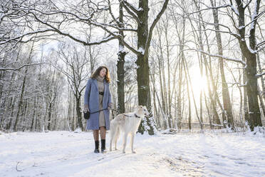 Glückliche Frau steht mit Windhund vor kahlen Bäumen im Winterpark - EYAF02486