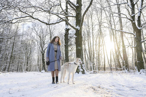 Happy woman standing with greyhound dog in front of bare trees at winter park - EYAF02486