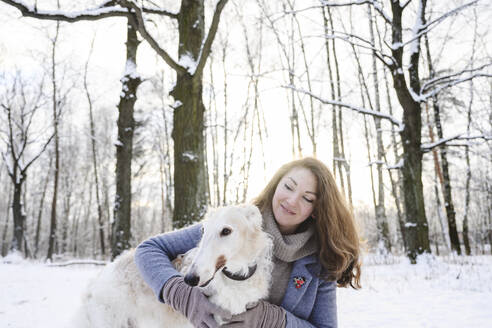 Smiling mature woman embracing greyhound dog in winter park - EYAF02487