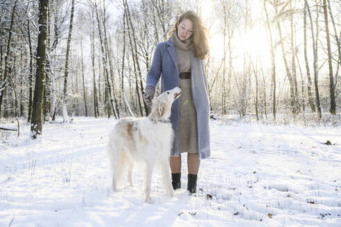 Ältere Frau mit Windhund im Schnee im Wald stehend - EYAF02489