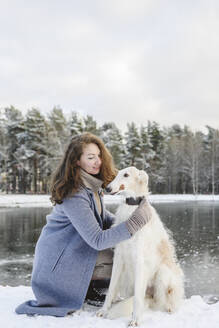 Mature woman stroking greyhound dog in front of frozen lake - EYAF02495