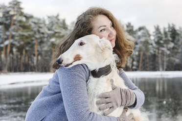 Reife Frau, die einen Windhund vor einem zugefrorenen See umarmt - EYAF02496