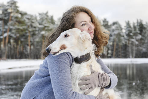 Reife Frau, die einen Windhund vor einem zugefrorenen See umarmt - EYAF02496
