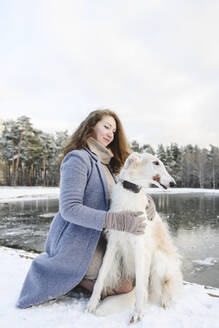 Woman stroking greyhound dog in front of frozen lake - EYAF02497