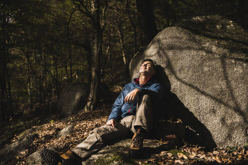 Älterer Mann lehnt an einem Felsen mit geschlossenen Augen im Wald - UUF27836