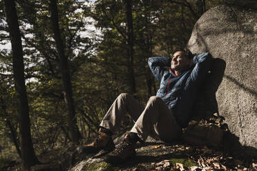 Mann mit Händen hinter dem Kopf entspannt sich an einem Felsen im Wald - UUF27840