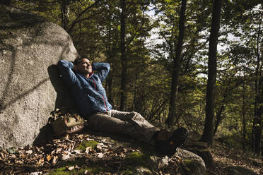 Älterer Mann mit Händen hinter dem Kopf, der sich an einem Felsen im Wald entspannt - UUF27841