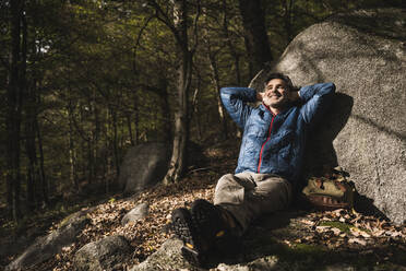 Glücklicher reifer Mann mit Händen hinter dem Kopf, der sich an einem Felsen im Wald entspannt - UUF27842