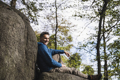 Lächelnder reifer Mann sitzt an einem Felsen vor Bäumen - UUF27843