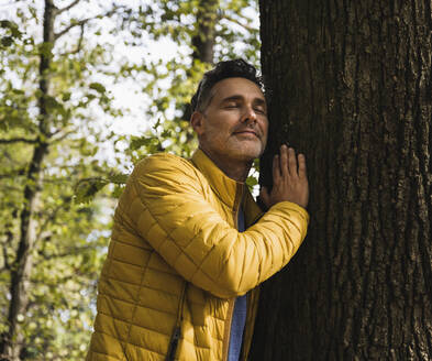 Lächelnder reifer Mann, der einen Baum im Wald umarmt - UUF27862