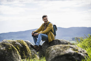 Glücklicher reifer Mann mit Rucksack sitzt auf einem Felsen vor dem Himmel - UUF27864