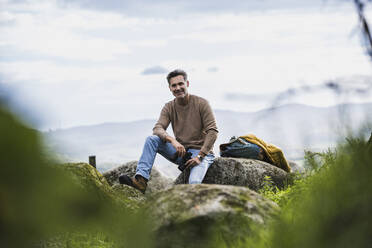 Glücklicher reifer Mann sitzt auf einem Felsen vor dem Himmel - UUF27865