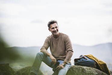 Happy mature man with binoculars sitting on rock - UUF27866