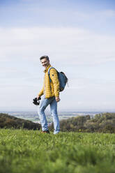 Happy mature man walking with backpack and binoculars under sky - UUF27868