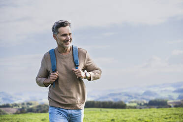 Smiling mature man with backpack walking in front of sky - UUF27879