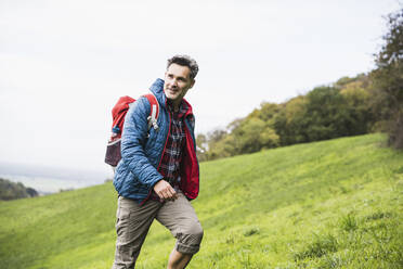 Lächelnder reifer Mann mit Rucksack auf Gras unter Himmel spazierend - UUF27891