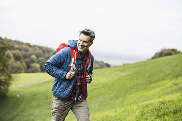 Lächelnder Mann mit Rucksack wandert auf Gras unter Himmel - UUF27892