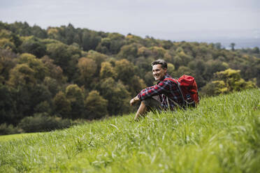 Lächelnder Mann mit Rucksack im Gras sitzend - UUF27909