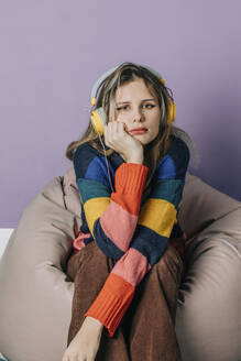 Sad girl listening to music through headphones sitting on bean bag against purple background - VSNF00276