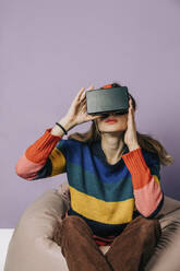 Mädchen, das einen Simulator der visuellen Realität trägt und auf einem Bohnensack vor einem lila Hintergrund sitzt - VSNF00278