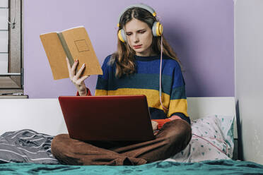 Girl using laptop doing homework sitting on bed in front of wall - VSNF00284
