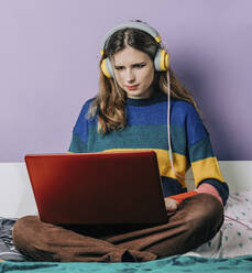 Mädchen mit Kopfhörern bei der Hausarbeit am Laptop vor einer lila Wand - VSNF00285