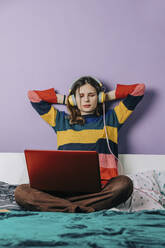 Mädchen mit Händen hinter dem Kopf mit Laptop und Kopfhörer auf dem Bett zu Hause - VSNF00286