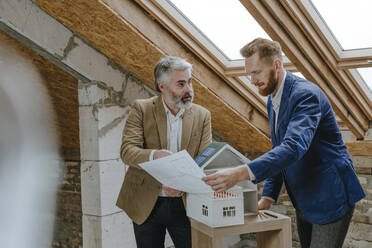 Architect explaining colleague over blueprint standing by house model at construction site - YTF00399