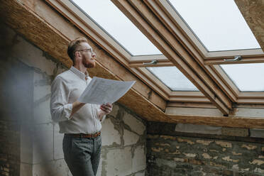 Thoughtful architect standing with blueprint looking through window at construction site - YTF00426