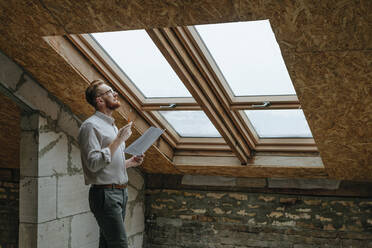 Contemplative architect with blueprint standing near window at home - YTF00428