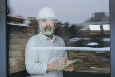 Thoughtful architect wearing hardhat standing with dairy seen through glass - YTF00432