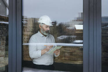 Businessman wearing hardhat making notes at construction site seen through glass - YTF00433