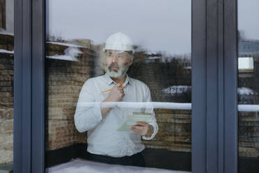 Contemplative businessman wearing hardhat holding diary seen through window glass - YTF00435