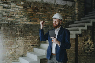 Happy architect with clenched fist holding tablet PC at construction site - YTF00440
