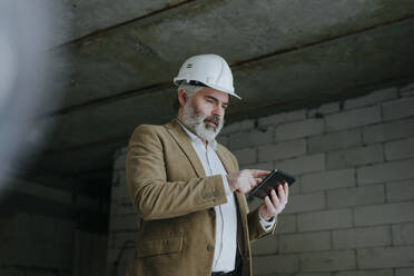 Architect wearing hardhat using tablet PC standing at construction site - YTF00443