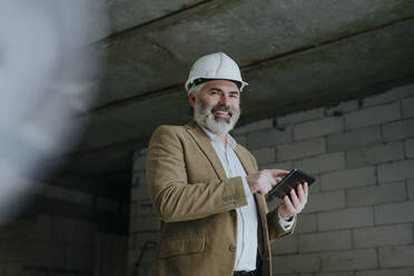 Happy businessman with hardhat standing with tablet PC at construction site - YTF00444