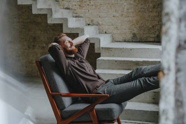 Happy businessman relaxing with hands behind head in chair at construction site - YTF00474