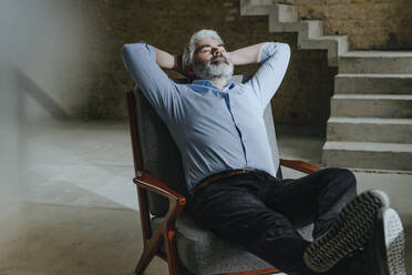 Mature architect relaxing in chair with hands behind head at under construction site - YTF00485