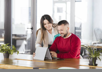 Smiling businesswoman discussing with colleague over tablet PC in coworking office - LJF02398