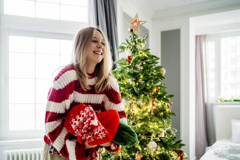 Fröhliches Mädchen hält Pullover am Weihnachtsbaum zu Hause - MDOF00470