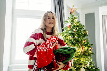 Lächelndes Mädchen mit einem Stapel von Pullovern und einem Weihnachtsbaum zu Hause - MDOF00471