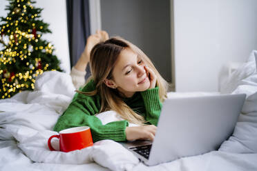 Lächelndes Mädchen schaut Film auf Laptop mit roter Tasse auf Bett zu Hause - MDOF00481