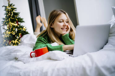 Glückliches Mädchen mit Laptop auf dem Bett mit Weihnachtsbaum im Hintergrund zu Hause - MDOF00482