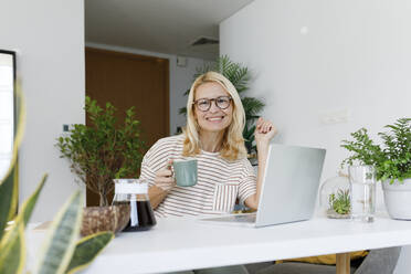 Smiling freelancer with tea cup and laptop sitting at desk - TYF00574